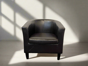 A brown pleather tub chair with a hard backrest and seat, standing against a white brick wall on a black textured floor.