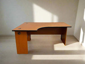 A small L-shaped desk with a beige laminate top and wooden legs, situated against a white brick wall on a concrete floor.