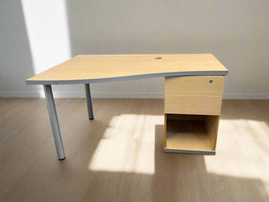 A wooden desk with a gray metal leg and a matching wooden creditena with a lower shelf, placed against a white brick wall on a black grate floor.