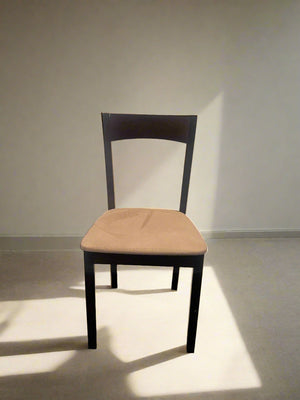 A dining chair with a dark wood frame and a cream seat, standing against a white brick wall on a grey floor.
