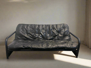 Black pleather sleeper couch with a steel frame, situated against a white brick wall on a concrete floor.