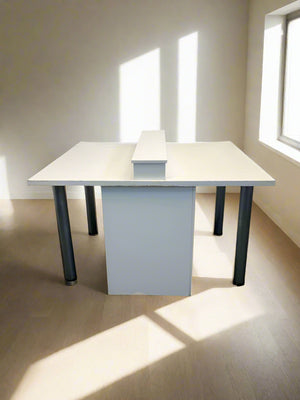 A white wooden multi-person desk with a divider, metal legs, and situated against a white brick wall.