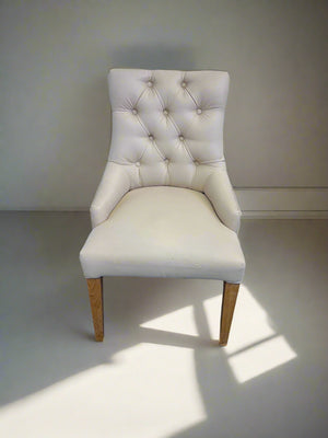 An elegant cream upholstered dining chair with button-tufted backrest and oak wooden legs, against a white brick background.