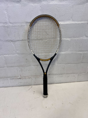 Tennis racket with black and gold frame against a light gray brick wall.