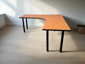 An L-shaped desk with an oak finish top and black metal legs, situated against a white brick wall on a grey floor.