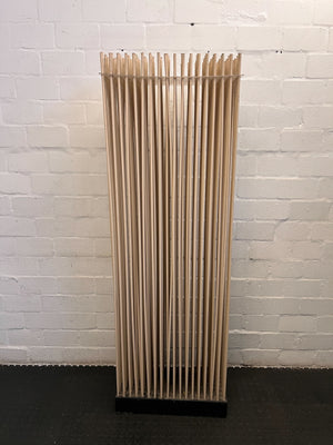 Tall, cylindrical wooden object with slotted design against a white brick wall.