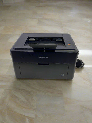 Samsung ML-1640 black laser printer with minor scratches, placed on a white surface against a brick wall background.
