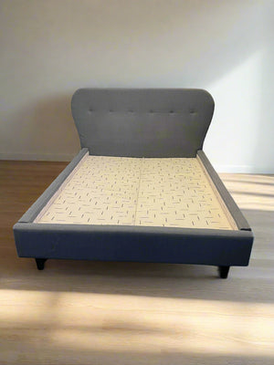 Gray upholstered bed with a wooden slatted base against a white brick wall.