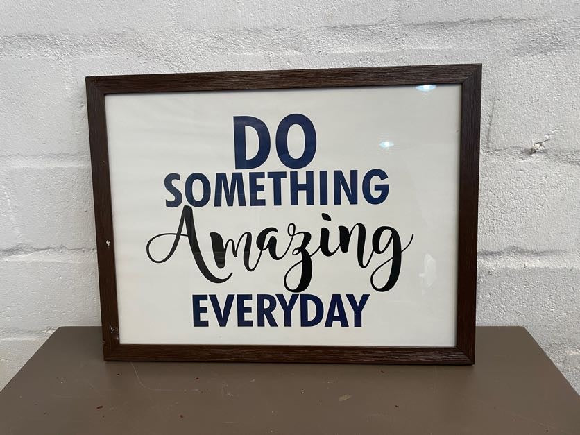 A framed artwork with the text 'DO SOMETHING AMAZING EVERYDAY' in a cursive script, set against a white background, housed in a brown wooden frame.