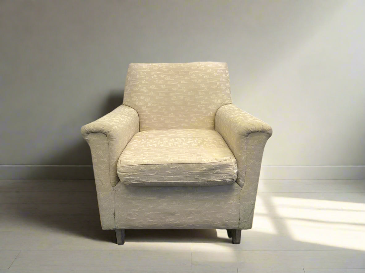 A cream fabric one seater armchair with a wooden frame, positioned against a white brick wall on a concrete floor.