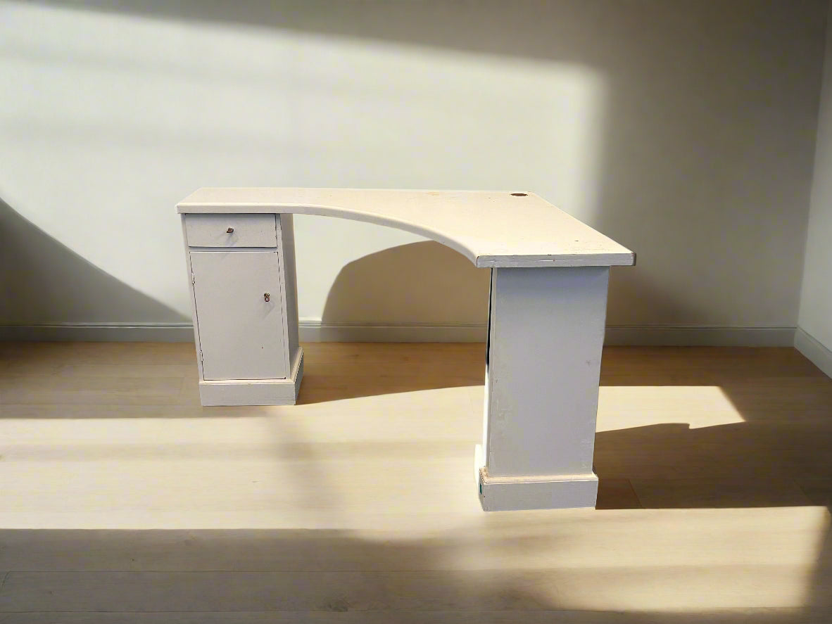 A white one drawer L-shaped desk with a modern design, placed against a brick wall on a black floor, with no visible drawers or visible brand markings.