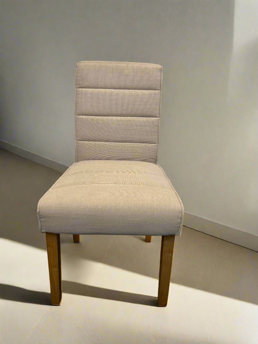 A beige upholstered dining chair with wooden legs against a white brick wall, with no other objects around.