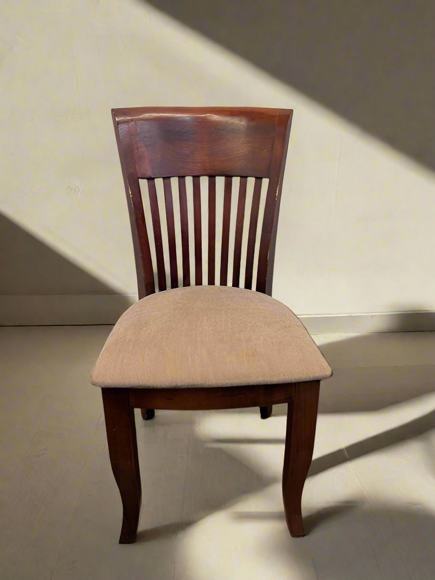 A single wooden dining room chair with a beige upholstered seat and a wooden backrest, positioned against a white brick wall on a patterned floor.