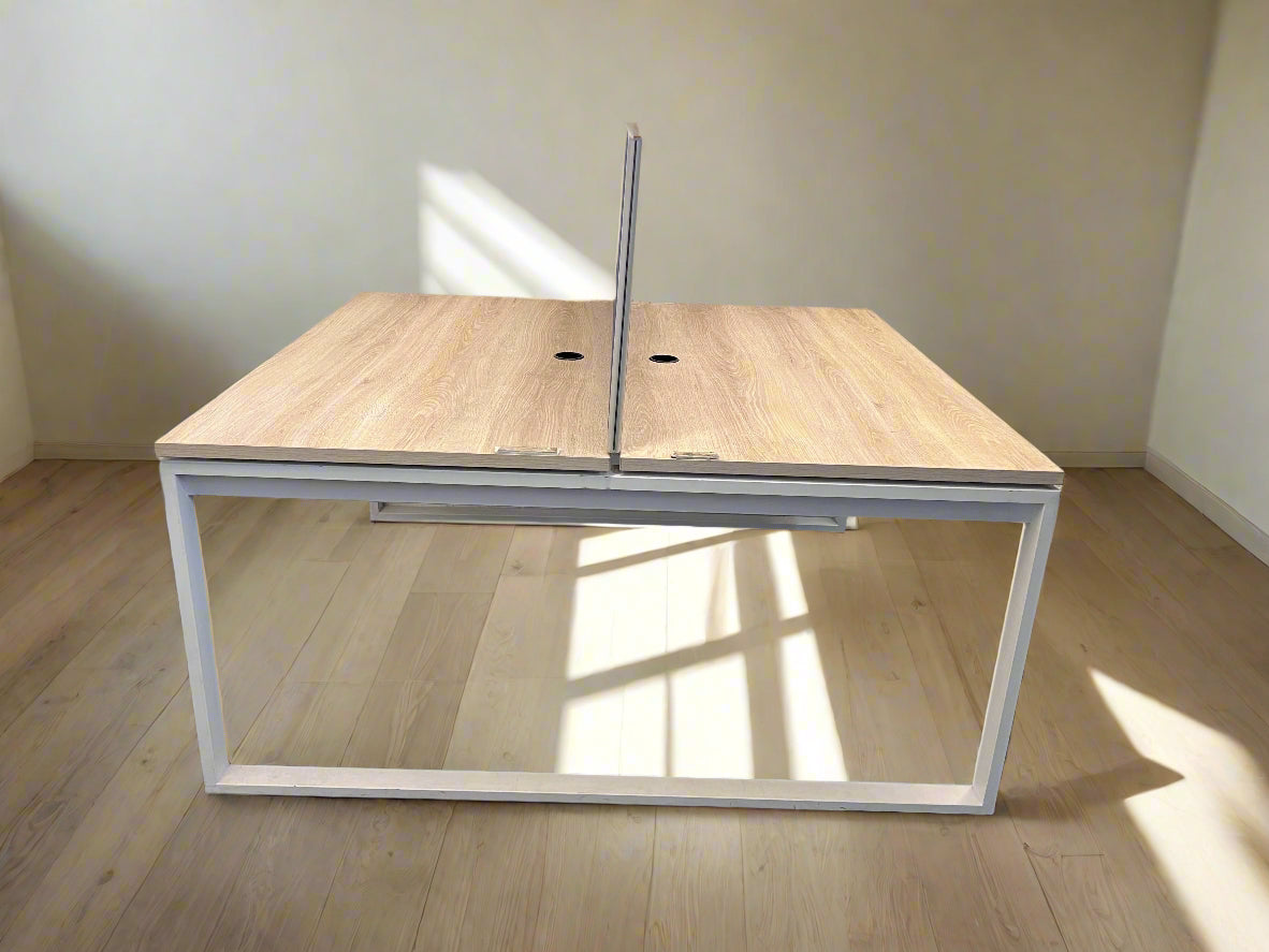 A contemporary split office desk with a white metal frame and light wood finish tabletop, viewed from an angle against a white brick wall.