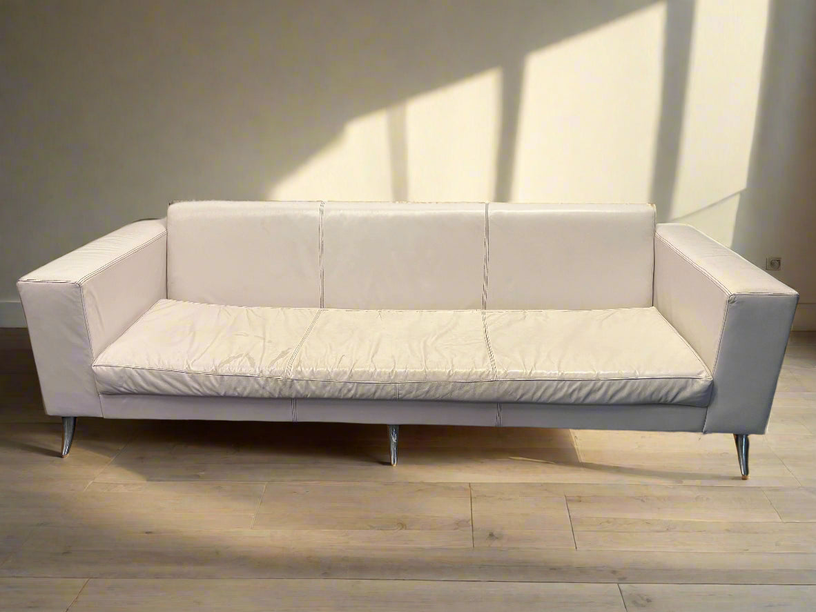 A white leather three-seater couch with minor wear, displayed against a white brick wall on a dark floor.