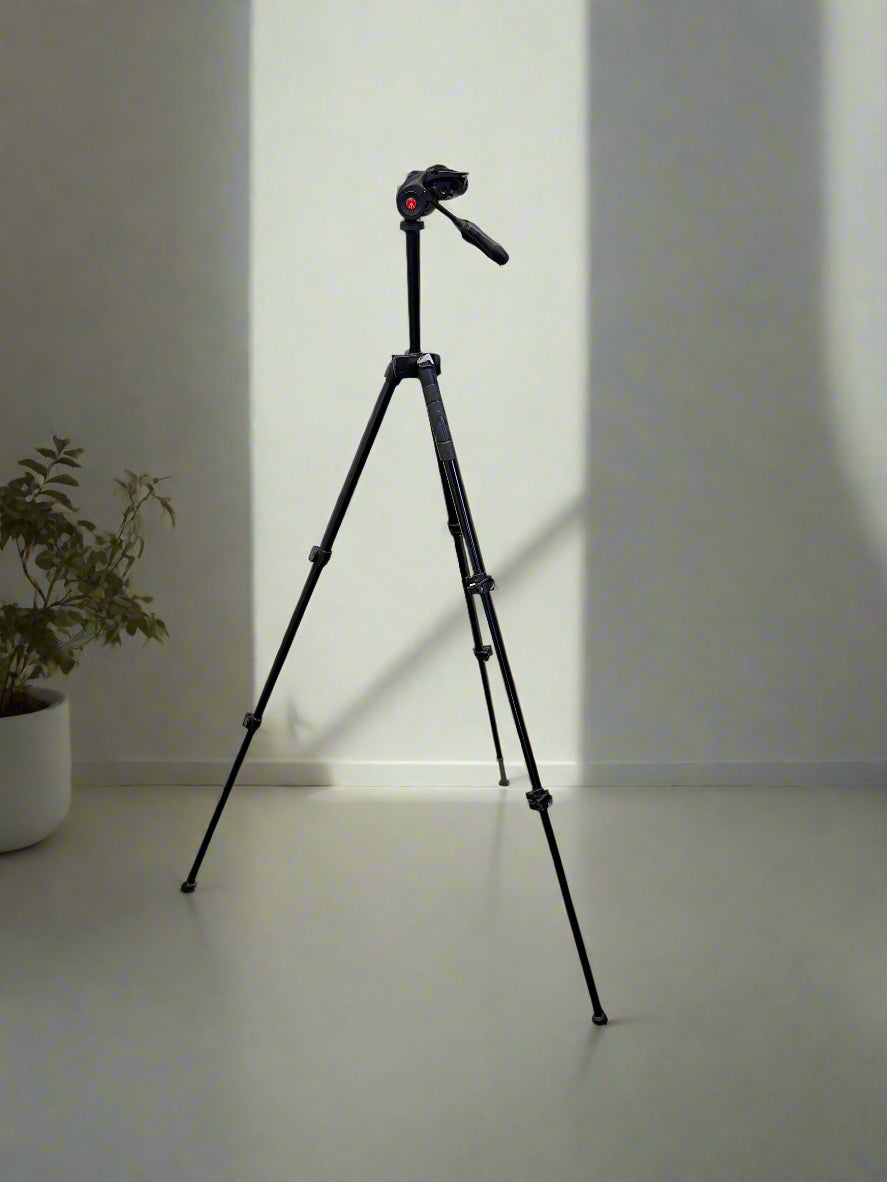 Camera on a tripod against a plain wall with a plant to the left.