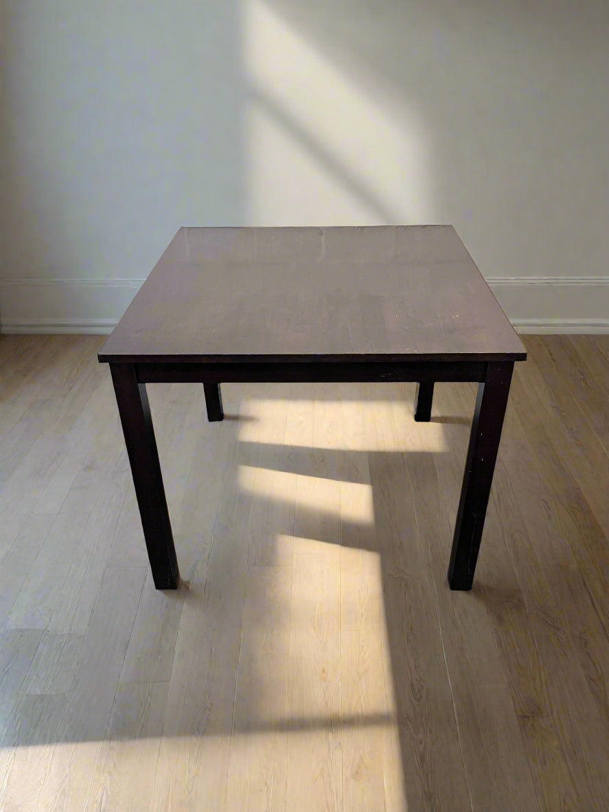 Square table with a dark finish on a wooden floor with light gray walls.