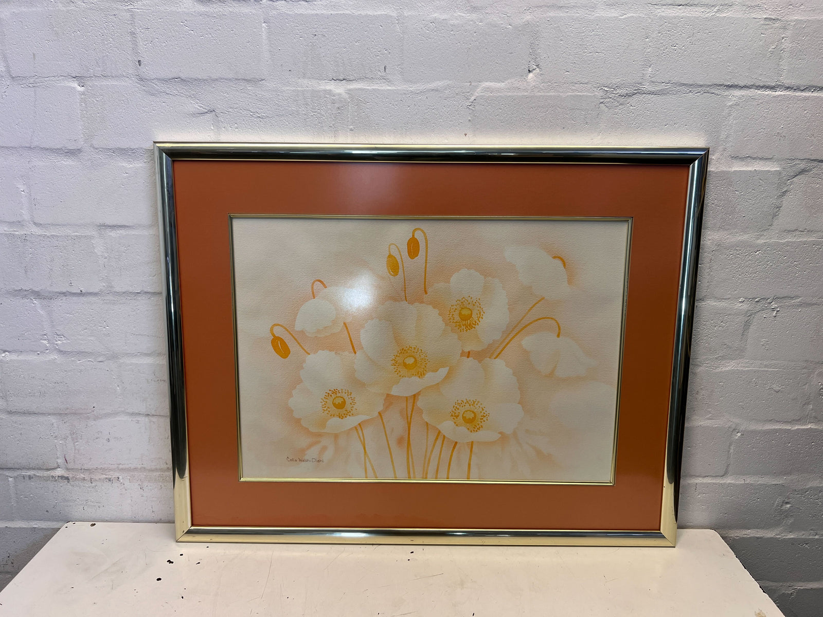 Framed artwork of white flowers with a gold border on a gray brick wall.