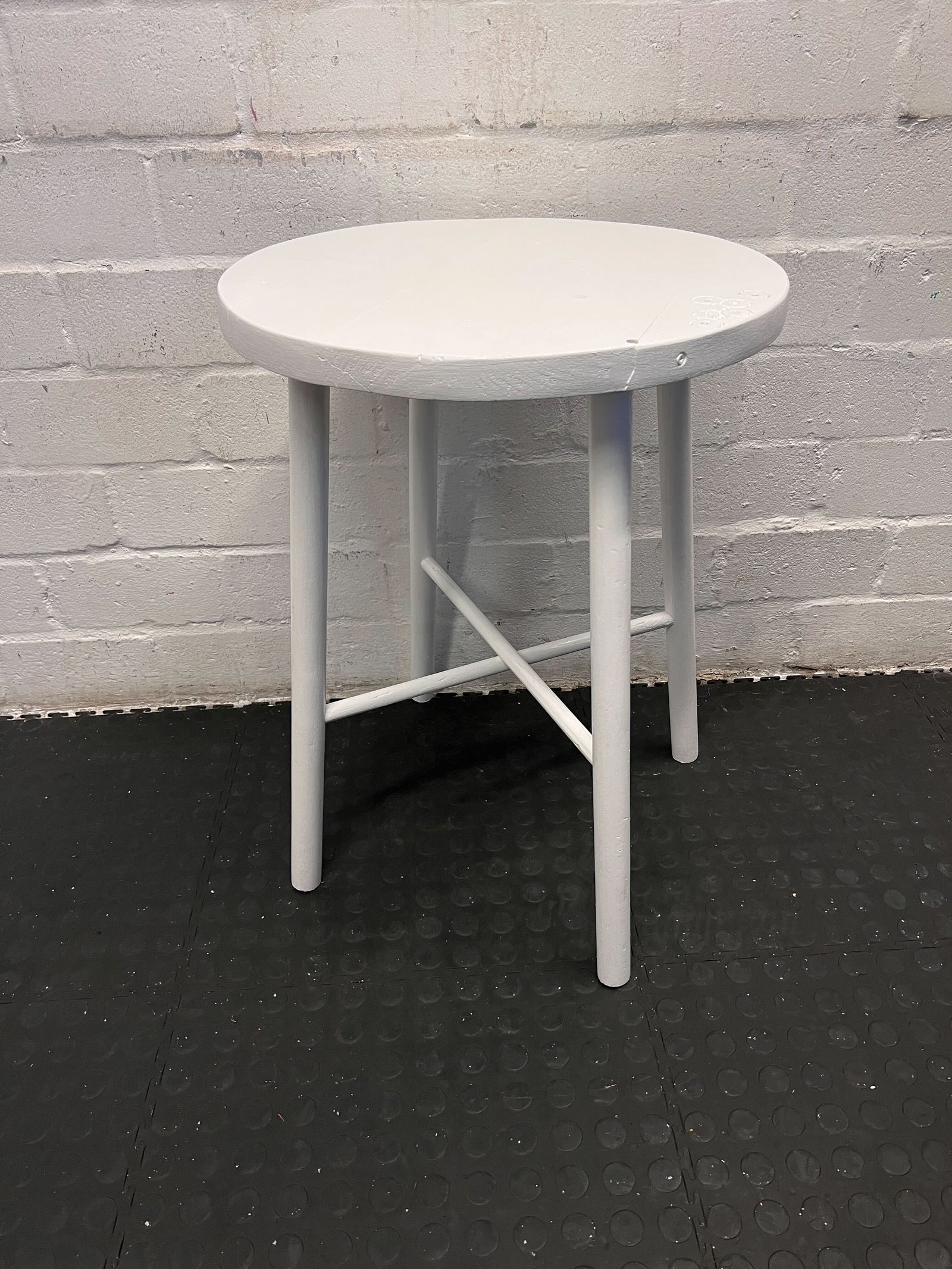 Round white table with four legs on a black floor against a gray brick wall.