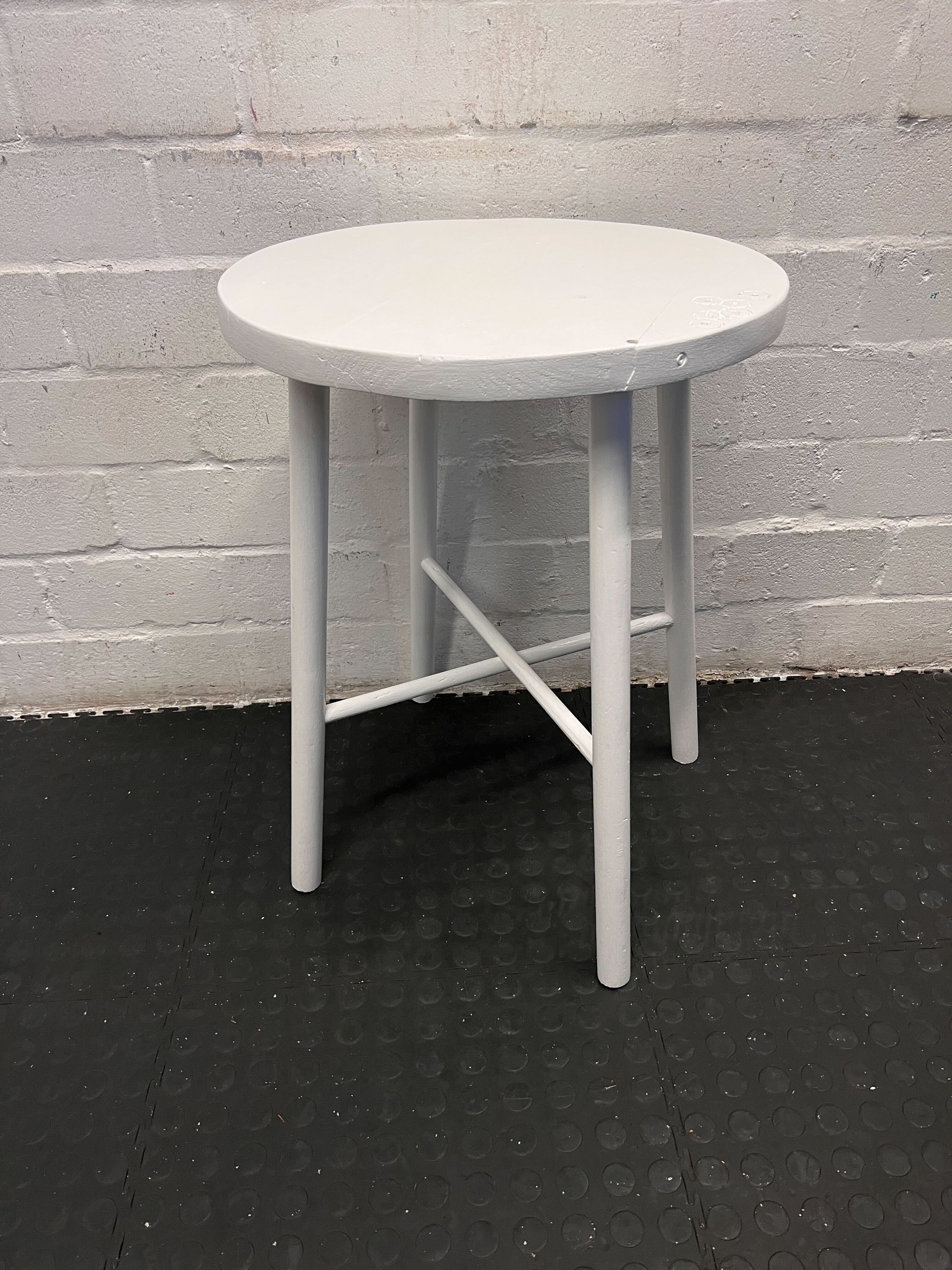 Round white table with four legs on a black floor against a gray brick wall.