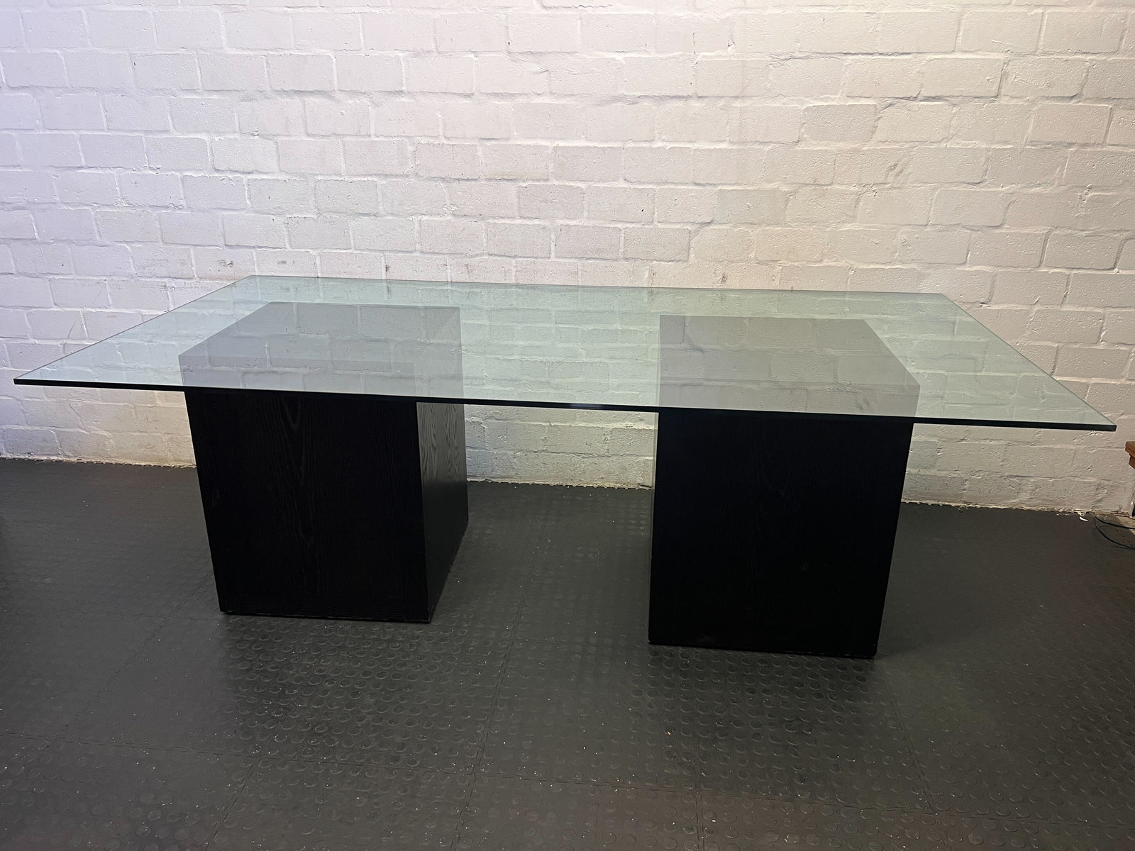 Glass dining table with black legs against a white brick wall.