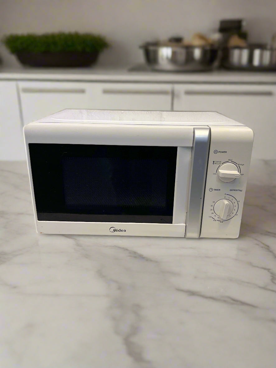 A compact white Midea microwave oven with minor scuffs on the exterior, placed on a table against a white brick wall background.