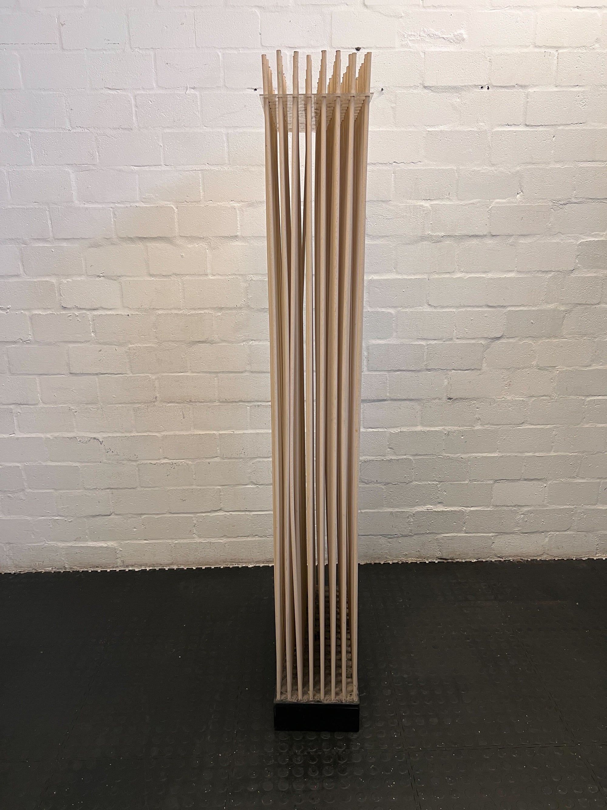 Tall, cylindrical wooden object with slits against a white brick wall.