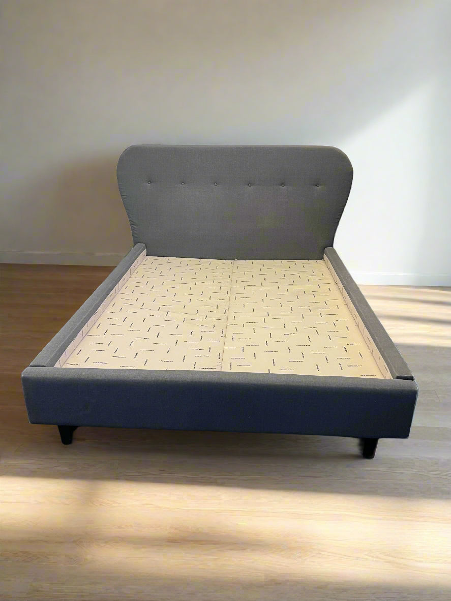 Gray upholstered bed with a wooden slatted base against a white brick wall.