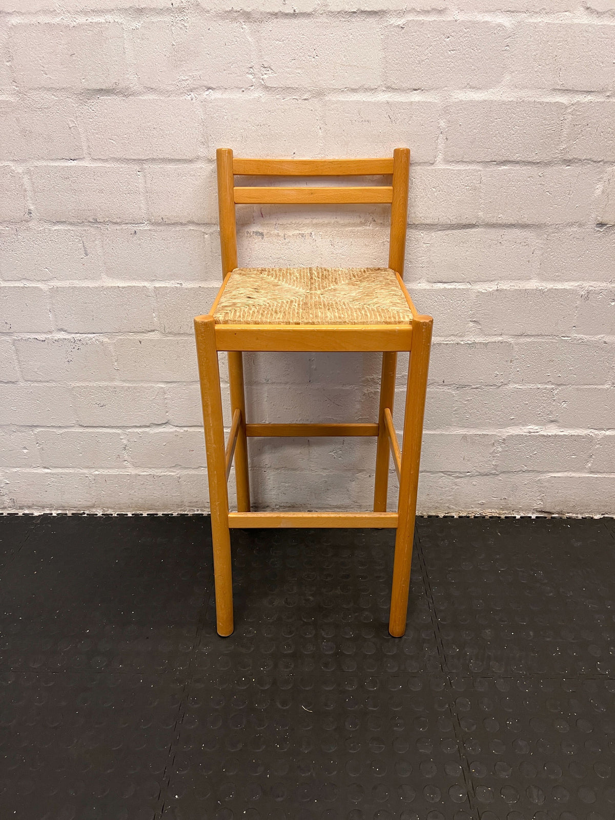 Wooden bar stool with a woven seat against a white brick wall.