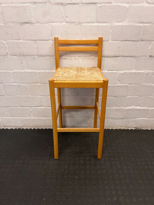 Wooden bar stool with a woven seat against a white brick wall.