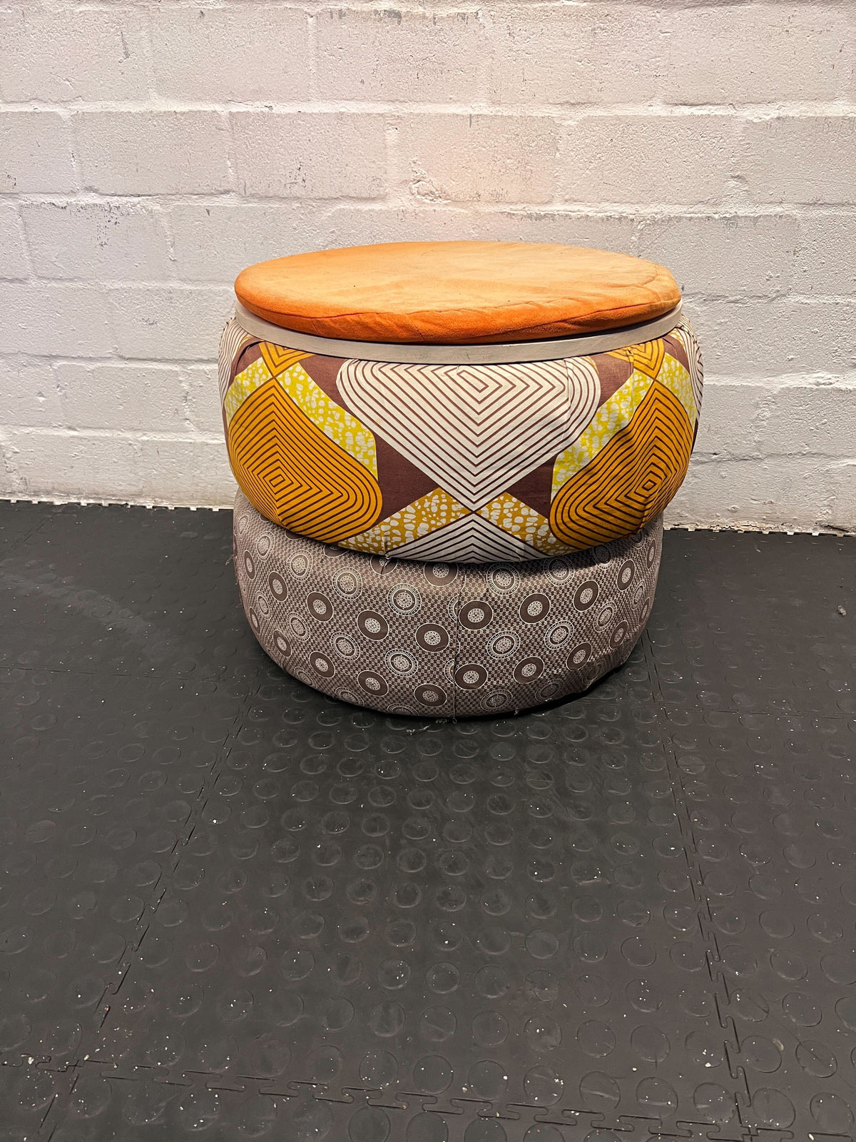 Two fabric-covered stools with patterned tops and a plain bottom, stacked against a gray brick wall.