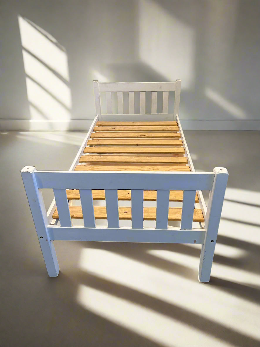 White wooden single bed frame with slats, positioned against a white brick wall on a black surface.